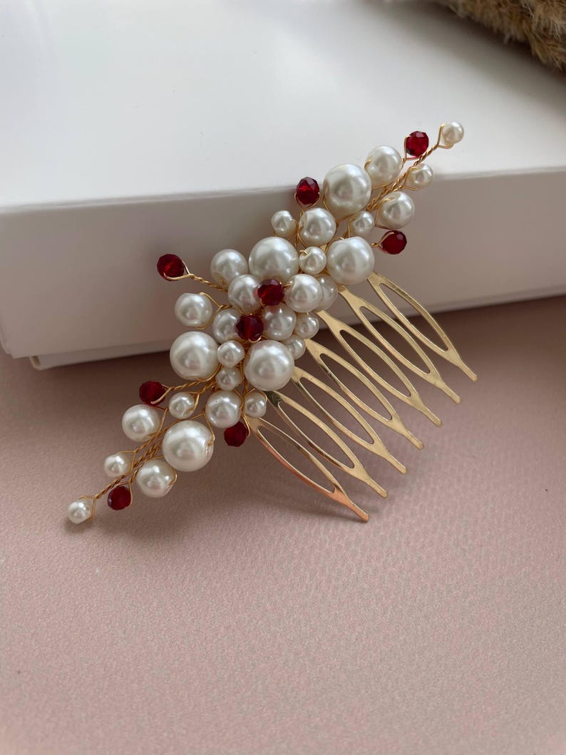 May include: A gold-toned hair comb adorned with clusters of faux pearls and red beads. The comb features a series of tines for securing in hair. The pearls vary in size, creating a textured, elegant look. The red beads add a pop of color.