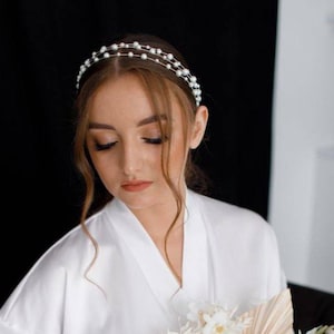 May include: A woman wearing a white satin robe with a pearl headband. She is holding a bouquet of white flowers.