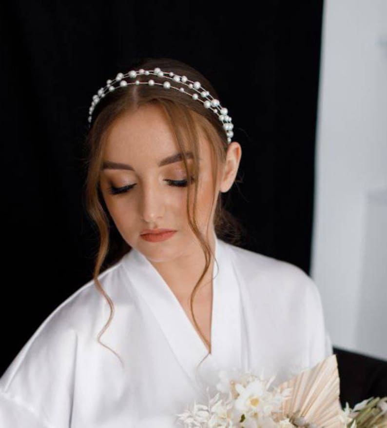 May include: A woman wearing a white satin robe with a pearl headband. She is holding a bouquet of white flowers.