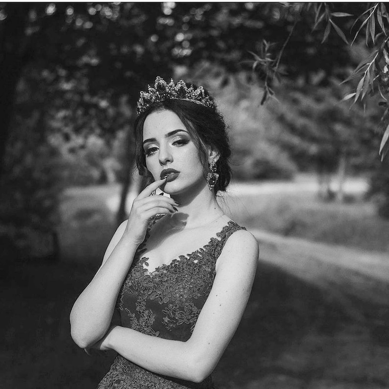 Puede incluir: Una mujer con un vestido de encaje negro y una corona de joyas, con la mano en la barbilla, mirando pensativamente a la c&aacute;mara.