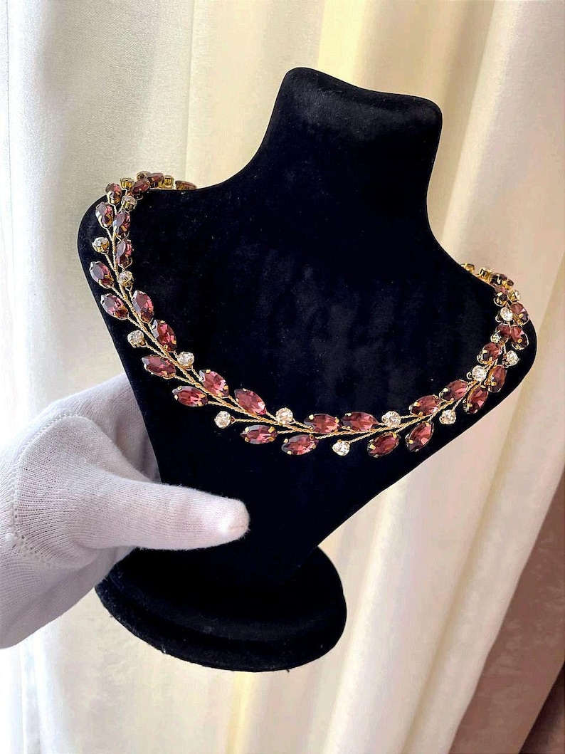 May include: A gold necklace with a delicate design featuring small, oval-shaped purple gemstones and tiny white rhinestones. The necklace is displayed on a black velvet mannequin.