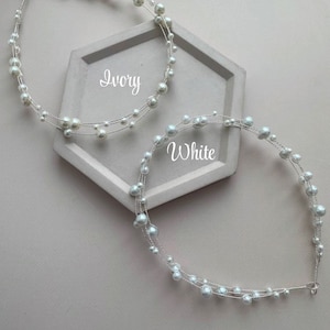 May include: Two pearl headbands, one in ivory and one in white, on a gray hexagonal surface. The headbands are made of wire with small pearls strung along it.