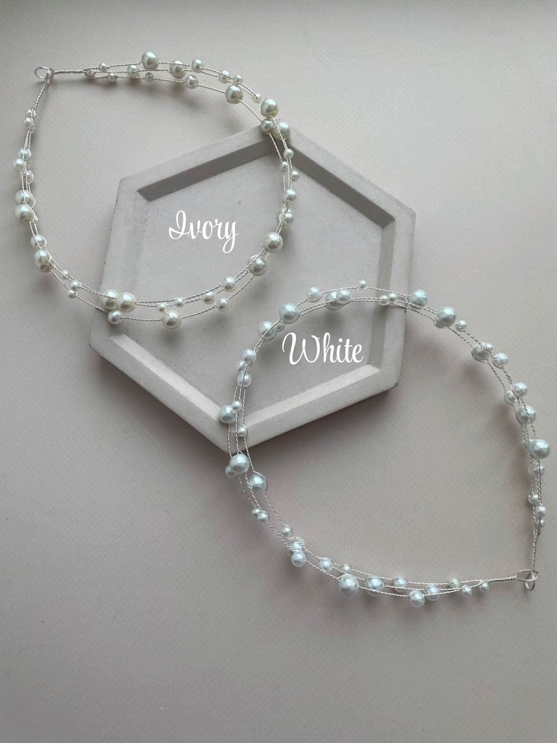 May include: Two pearl headbands, one in ivory and one in white, on a gray hexagonal surface. The headbands are made of wire with small pearls strung along it.