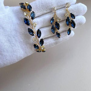 May include: A set of gold-toned jewellery featuring a bracelet and earrings. The bracelet and earrings are adorned with blue and clear rhinestones in a leaf-like design.