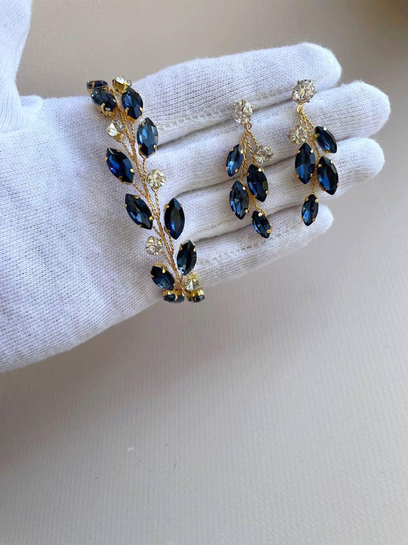 May include: A set of gold-toned jewellery featuring a bracelet and earrings. The bracelet and earrings are adorned with blue and clear rhinestones in a leaf-like design.