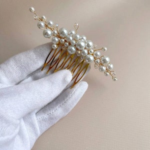 May include: A gold-toned hair comb adorned with clusters of white faux pearls and clear crystals. The comb features a series of fine, evenly spaced teeth. The pearls vary in size, creating a delicate, elegant design. This hair accessory is suitable for weddings or special occasions.