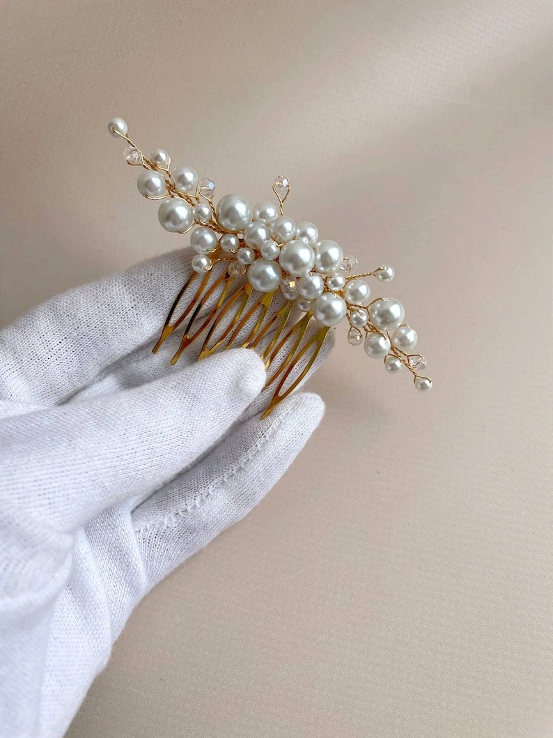May include: A gold-toned hair comb adorned with clusters of white faux pearls and clear crystals. The comb features a series of fine, evenly spaced teeth. The pearls vary in size, creating a delicate, elegant design. This hair accessory is suitable for weddings or special occasions.