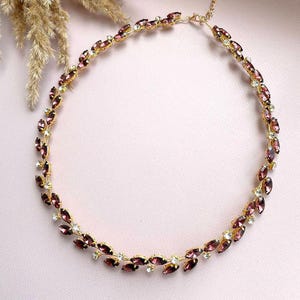 May include: A gold necklace with a delicate chain and a design of small, purple marquise-shaped rhinestones. The rhinestones are set in a leaf-like pattern.