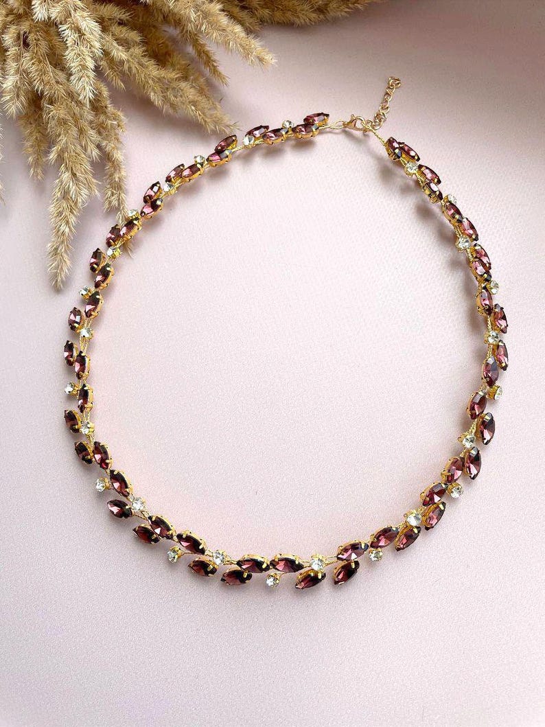 May include: A gold necklace with a delicate chain and a design of small, purple marquise-shaped rhinestones. The rhinestones are set in a leaf-like pattern.