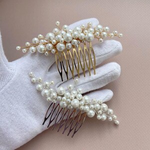 May include: Two pearl hair combs. Each comb features a cluster of white pearls attached to a gold-colored metal comb. The pearls vary in size, creating a textured, elegant look. The combs are designed for bridal or formal hairstyles.
