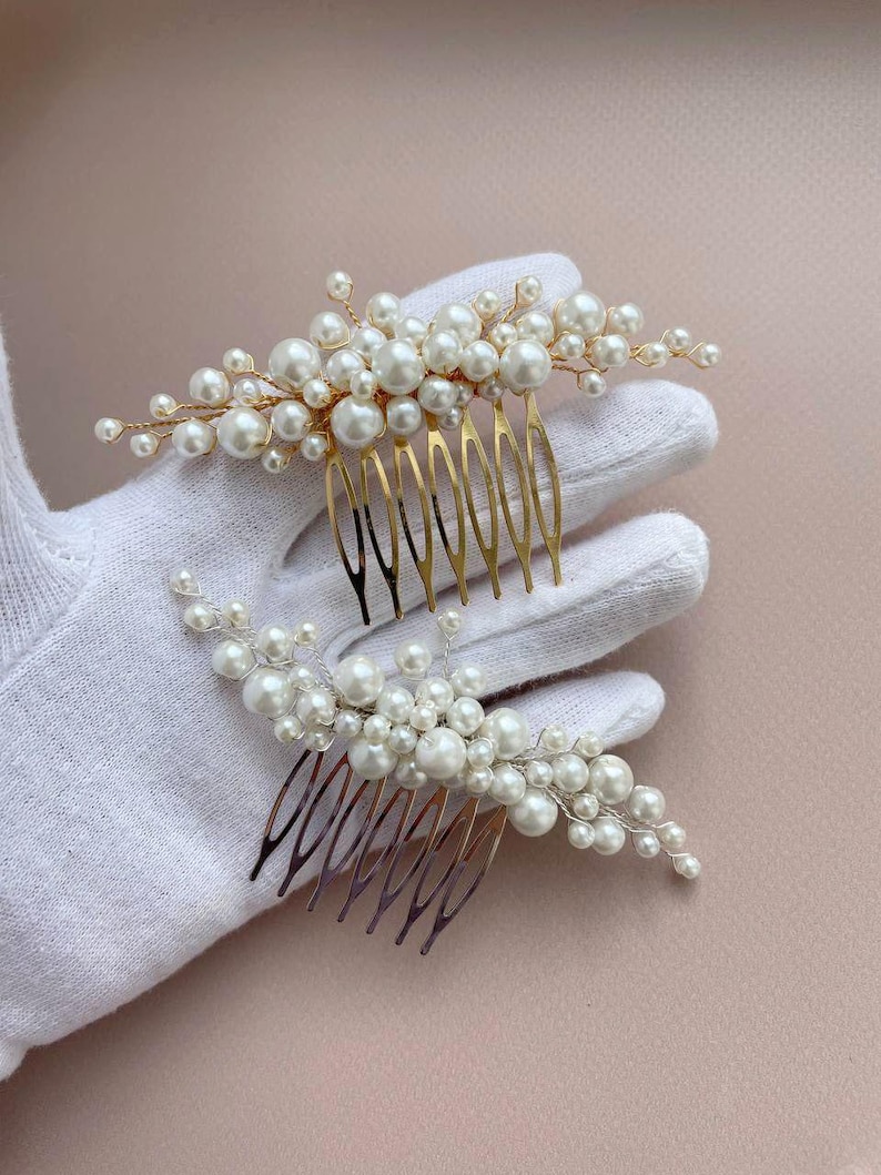 May include: Two pearl hair combs. Each comb features a cluster of white pearls attached to a gold-colored metal comb. The pearls vary in size, creating a textured, elegant look. The combs are designed for bridal or formal hairstyles.