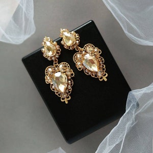 Puede incluir: Un par de pendientes colgantes de tono dorado con cristales en forma de l&aacute;grima. Los pendientes tienen un delicado dise&ntilde;o de filigrana.