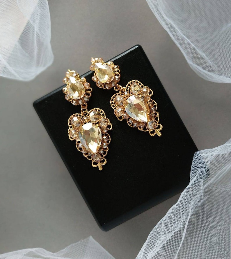 Puede incluir: Un par de pendientes colgantes de tono dorado con cristales en forma de l&aacute;grima. Los pendientes tienen un delicado dise&ntilde;o de filigrana.