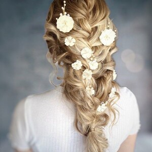 以下が含まれることがあります： 白い花のヘアクリップで飾られたサイドブレードのブロンドの女性。花はブレードに沿ってカスケード状に配置されています。