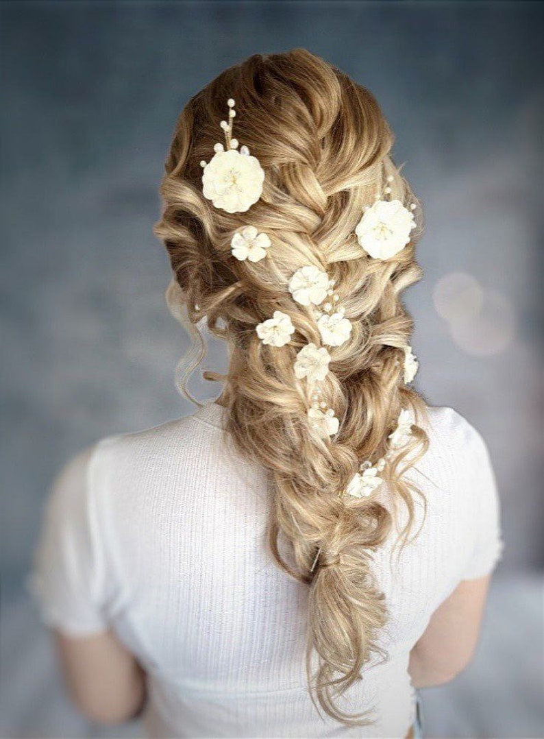 以下が含まれることがあります： 白い花のヘアクリップで飾られたサイドブレードのブロンドの女性。花はブレードに沿ってカスケード状に配置されています。