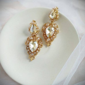 Puede incluir: Un par de pendientes colgantes de tono dorado con cristales transparentes en forma de l&aacute;grima. Los pendientes tienen un dise&ntilde;o delicado e intrincado.
