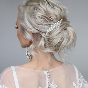 May include: A woman's hairstyle with light blonde, wavy hair styled in a low bun. A pearl hair clip is placed on the side. The woman is wearing a white dress with a sheer back and pearl buttons.