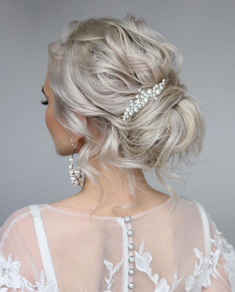 May include: A woman's hairstyle with light blonde, wavy hair styled in a low bun. A pearl hair clip is placed on the side. The woman is wearing a white dress with a sheer back and pearl buttons.