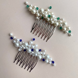 May include: Two decorative hair combs. One features white pearls and green beads, the other white pearls and blue beads. Both have silver-colored metal combs. The combs are arranged on a light pink surface.
