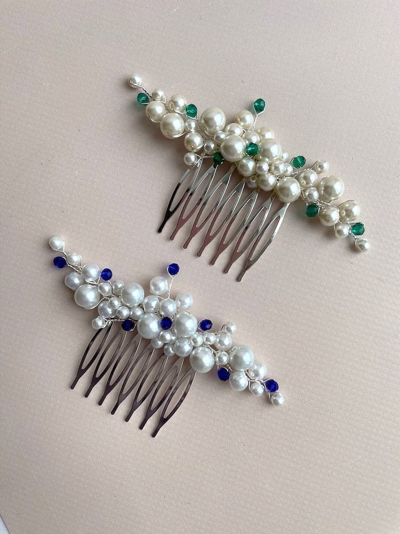 May include: Two decorative hair combs. One features white pearls and green beads, the other white pearls and blue beads. Both have silver-colored metal combs. The combs are arranged on a light pink surface.