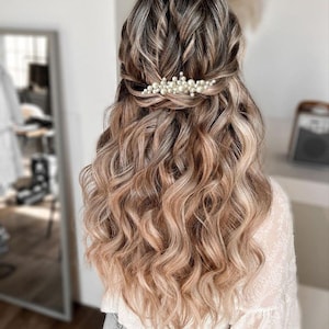 May include: A woman's hairstyle featuring long, wavy, light brown hair with blonde highlights. A pearl hair clip adorns the back of the head, holding a portion of the hair in place. The hair cascades down the back.