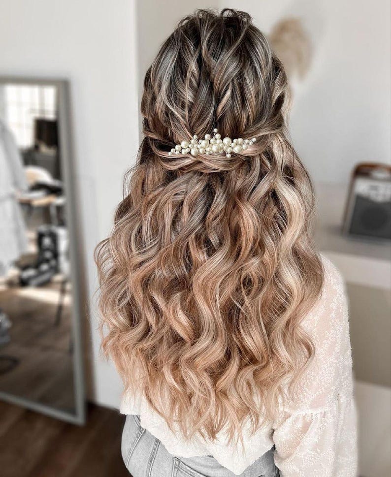 May include: A woman's hairstyle featuring long, wavy, light brown hair with blonde highlights. A pearl hair clip adorns the back of the head, holding a portion of the hair in place. The hair cascades down the back.