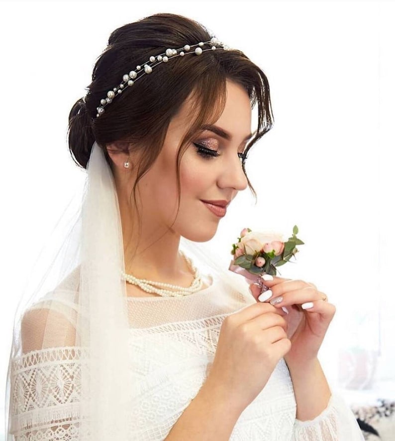 May include: A bride wearing a white lace dress with a veil and a pearl headband. She is holding a small bouquet of pink roses.