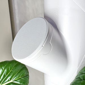 May include: A close-up of a white, angled, cylindrical object with a textured, circular top. The object has a curved design and is positioned near green leafy plants. The background is a light color.