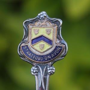 May include: A silver spoon with a decorative handle and a shield-shaped emblem. The emblem features a yellow and blue design with the words "STRATFORD-ON-AVON" in a blue banner. The background is a blurred green.