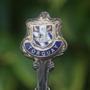 May include: Silver souvenir spoon with a blue and white enamel crest featuring a coat of arms and the word "TORQUAY" in gold lettering.