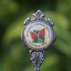 May include: A silver spoon with a round, white enamel charm featuring a butterfly and the text "National Garden Festival Gateshead 1990".
