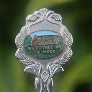 Könnte beinhalten: Ein silberner Löffel mit einem verzierten Griff und einem dekorativen Kopf. Der Kopf zeigt ein ovales Design mit einer Landschaftsszene und dem Text "WIDECOMBE IN THE MOOR". Die Szene umfasst Gebäude und einen blauen Himmel.