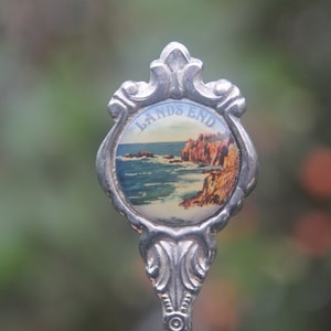 May include: Silver souvenir spoon with a blue and green image of a coastal scene and the text "Land's End".