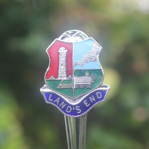 May include: A silver souvenir spoon with a blue, green, red, and white enamel design. The spoon features a shield with a lighthouse, a house, and a map. The text "LAND'S END" is written on the spoon.