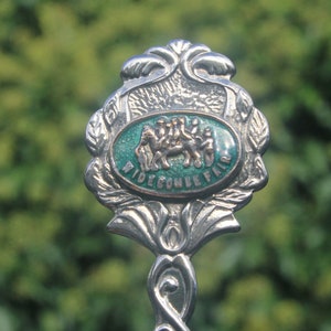 Könnte beinhalten: Ein silberner Souvenir-Löffel mit einem grünen Emaille-Design, das eine Gruppe von Menschen zeigt, die auf einem Kamel reiten. Der Löffel hat einen dekorativen Griff und die Worte "Ridecombe Fair" sind in das Emaille eingeprägt.