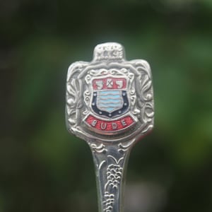 May include: Silver souvenir spoon with an ornate design featuring a shield with the text "BUD" and a blue and white crest. The shield is surrounded by a red and white border.