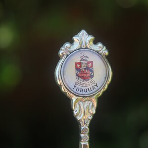 May include: A silver souvenir spoon with a white enamel design featuring a coat of arms and the text "Torquay".