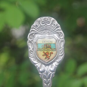 May include: Silver souvenir spoon with an ornate design and a shield featuring the word "DEVON" and a small image of a ship.