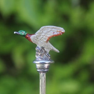 May include: A metal cocktail pick featuring a small, hand-painted mallard duck figurine at the top. The duck has a green head, a brown chest, and white wings with orange accents, mounted on a silver-toned base atop a thin, vertical metal stem.