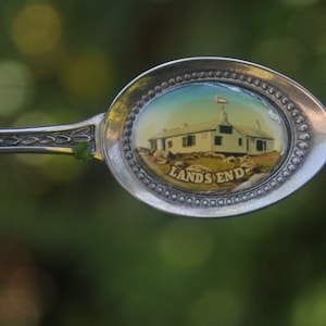 May include: A vintage silver spoon with an oval-shaped bowl featuring a painted image of a white building with a red and white flag, labeled "LANDS END". The spoon's handle has an ornate, decorative design.
