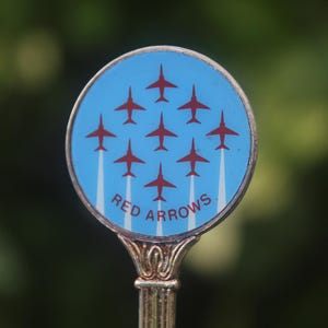 Może przedstawiać: Srebrzysta metalowa łyżeczka z okrągłym emblematem na górze. Emblemat ma jasnoniebieskie tło z dziewięcioma małymi czerwonymi sylwetkami samolotów ułożonymi w romb, każda z białą smugą dymu. Napis RED ARROWS jest wydrukowany na czerwono u dołu koła.