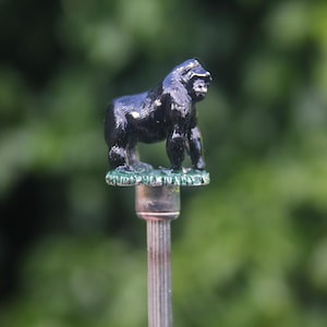 May include: A small, black gorilla figurine atop a silver-colored metal rod. The gorilla is standing on a green base. The background is a blurred green.