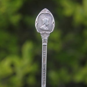 Op de afbeelding: Een zilveren lepel met een ovale kop met een gekroond profiel. De steel is gegraveerd met "CORONATION 1953". De lepel heeft een sierlijk ontwerp en staat tegen een vage groene achtergrond.