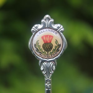 Op de afbeelding: Een zilveren souvenirlepel met een decoratief embleem. Het embleem toont de tekst "CALLANDER SCOTLAND" en een distel in rood, groen en geel. De lepel staat tegen een vage groene achtergrond. De distel is een nationaal symbool van Schotland.