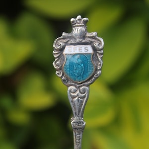May include: A silver spoon with a decorative handle and a shield-shaped emblem. The emblem features a blue enamel background with a white border and the word "EPES". A crown sits atop the emblem.