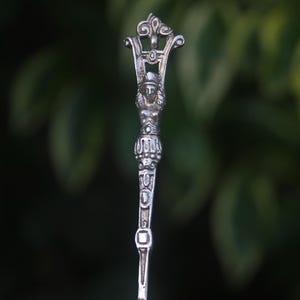 May include: A silver-toned decorative spoon with an ornate handle. The handle features a sculpted female figure with a crown, and detailed embellishments. The spoon's design is reminiscent of classical art and is set against a blurred green background.