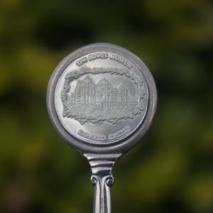 May include: A silver spoon with a circular emblem depicting a building and the words "THE BOWES MUSEUM" and "BARNARD CASTLE". The spoon has an ornate handle and is set against a blurred green backdrop.