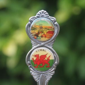 Può includere: Un manico di cucchiaio souvenir in metallo color argento con due inserti circolari. L'inserto superiore raffigura il sito minerario di Big Pit Blaenavon, mentre quello inferiore mostra il drago gallese rosso su sfondo verde e bianco con la scritta CYMRU.