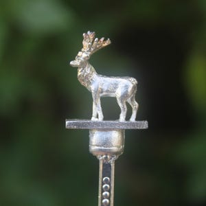 May include: A silver-coloured metal stirrer featuring a small, detailed figurine of a standing stag with antlers at the top. The deer is mounted on a flat rectangular base, which sits atop a cylindrical connector and a long, slender vertical stem.