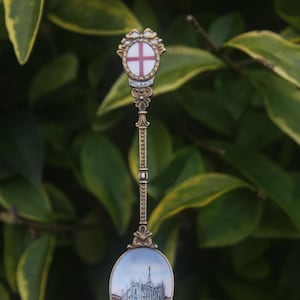 May include: A decorative souvenir spoon with a detailed design. The spoon features a crest with a red cross on a white background, and the word "MILANO". The bowl of the spoon has a painted image of a building. The spoon is set against a backdrop of green leaves.